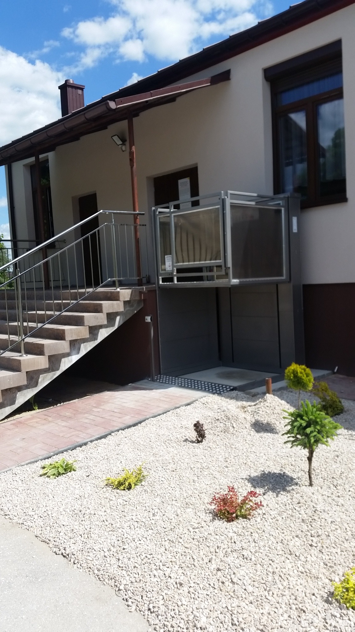 Zewnętrzna platforma schodowa dla osób niepełnosprawnych, zamontowana przy budynku z jasną elewacją, obok schodów z metalową barierką, otoczona ozdobnym, białym żwirem i niską roślinnością.