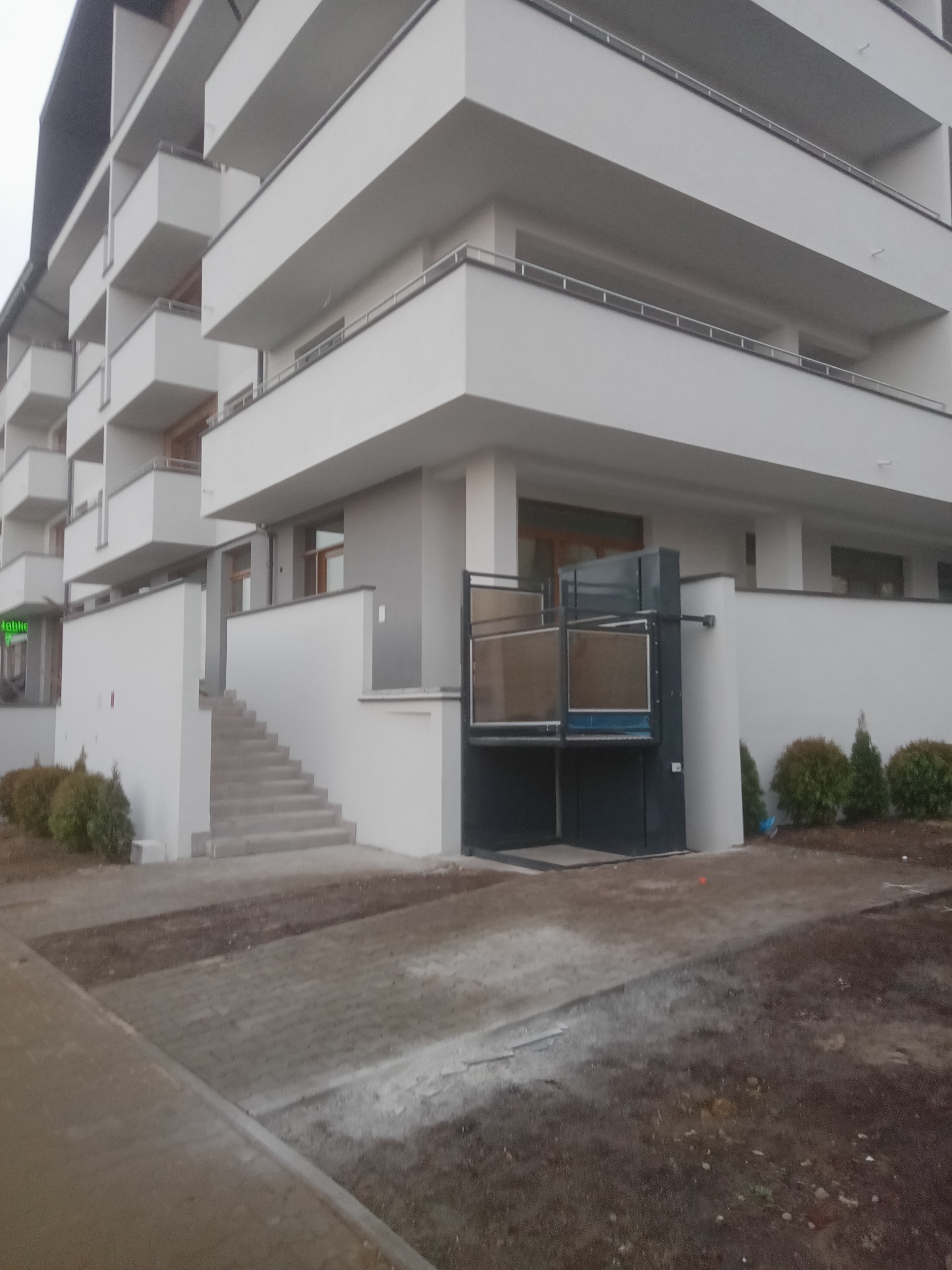 Zewnętrzna platforma schodowa dla osób z niepełnosprawnościami, zamontowana przy nowoczesnym budynku mieszkalnym z balkonami, widoczna z poziomu chodnika.