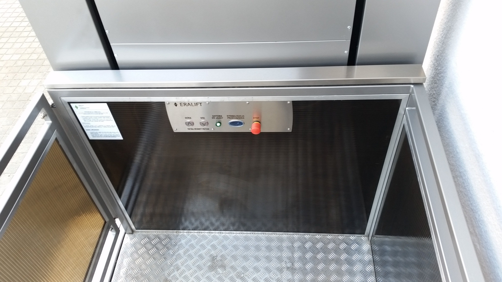 Wnętrze platformy schodowej dla osób niepełnosprawnych marki Eralift, widok z góry na panel sterowania z przyciskami i podłogę z blachy ryflowanej.