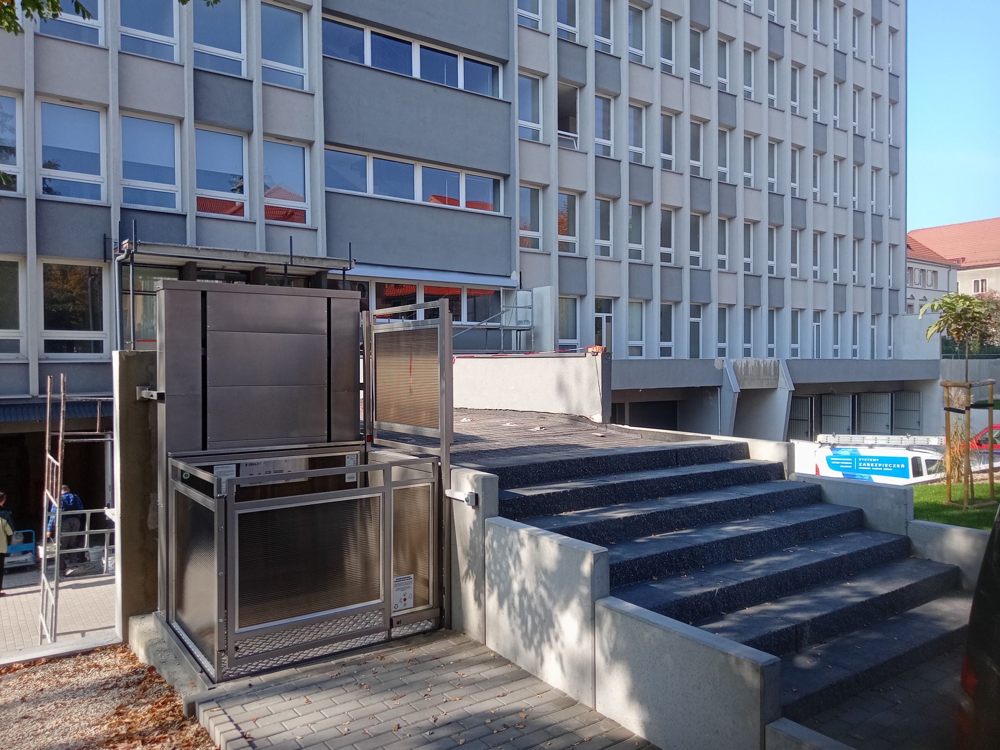 Zewnętrzna platforma schodowa dla osób niepełnosprawnych, zamontowana przy schodach budynku wielorodzinnego, z widoczną metalową konstrukcją i szybą ochronną.