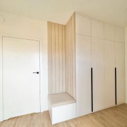 GONEK PIOTR - Biała szafa wnękowa z długimi, czarnymi uchwytami, zintegrowana z siedziskiem obitym beżową tkaniną i panelem ściennym w pionowe pasy, przy białych drzwiach.