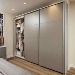 GONEK PIOTR - Nowoczesna szafa wnękowa z przesuwnymi, szarymi drzwiami w sypialni z drewnianą podłogą; otwarta część szafy z ubraniami na wieszakach i półkach.
