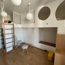 GONEK PIOTR - Pokój dziecięcy z zabudową na wymiar: antresola z barierką, schody z półkami, białe ściany z okrągłymi oknami, drewniana podłoga, oświetlenie punktowe, krzesło na kółkach i pufa.