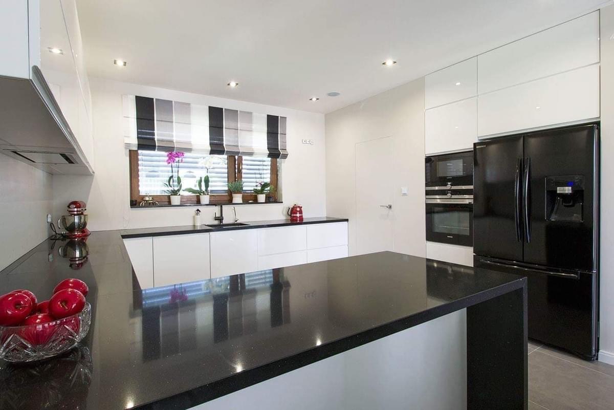 Nowoczesna kuchnia z białymi szafkami i czarnym blatem, okno z roletami w paski, czarna lodówka i piekarnik w zabudowie, owoce w szklanej misie na blacie.