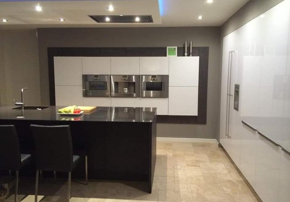 Nowoczesna kuchnia z białymi szafkami, wbudowanymi piekarnikami i czarnym, błyszczącym blatem wyspowym z przygotowanymi składnikami do sałatki, oświetlona punktowymi lampami sufitowymi.