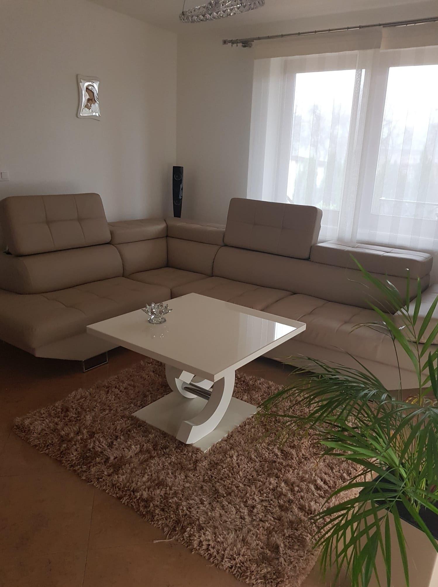 Beżowy narożnik z podnoszonymi zagłówkami w jasnym salonie, biały stolik kawowy z dekoracją, puszysty dywan i roślina doniczkowa.