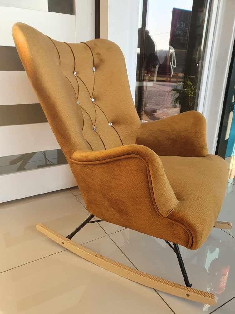 Złoty fotel bujany z tapicerowanym oparciem zdobionym kryształkami, na czarnych metalowych nogach i drewnianych płozach, ustawiony na jasnych płytkach.