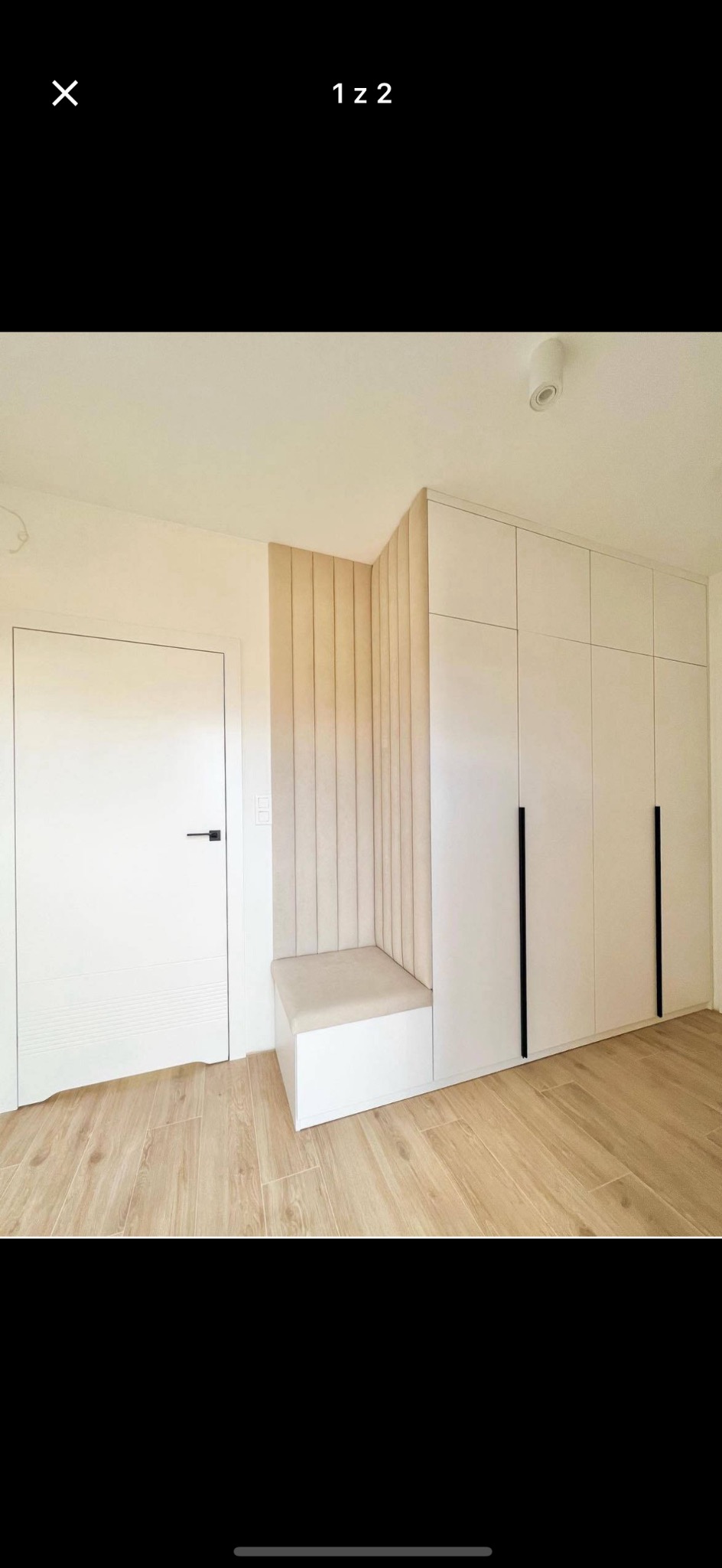 Biała szafa wnękowa z długimi, czarnymi uchwytami, zintegrowana z siedziskiem obitym beżową tkaniną i panelem ściennym w pionowe pasy, przy białych drzwiach.