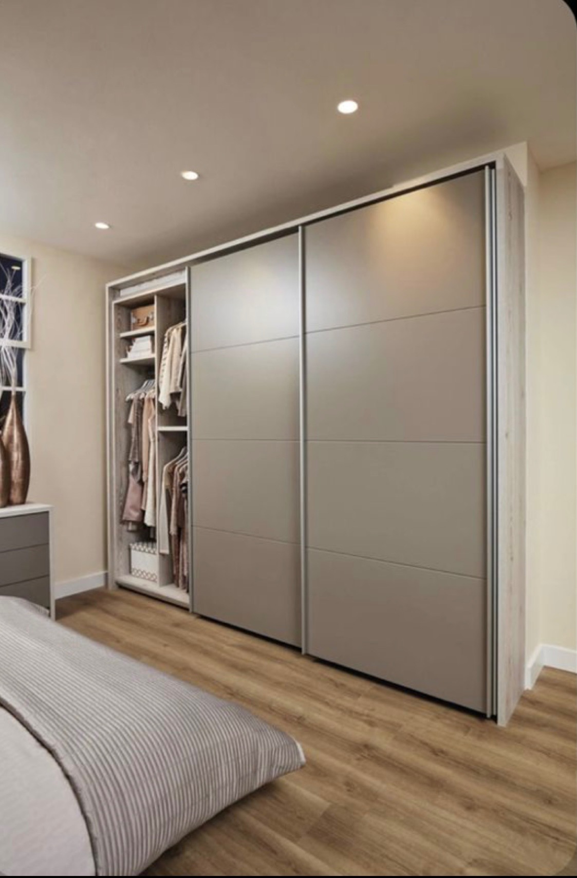 Nowoczesna szafa wnękowa z przesuwnymi, szarymi drzwiami w sypialni z drewnianą podłogą; otwarta część szafy z ubraniami na wieszakach i półkach.