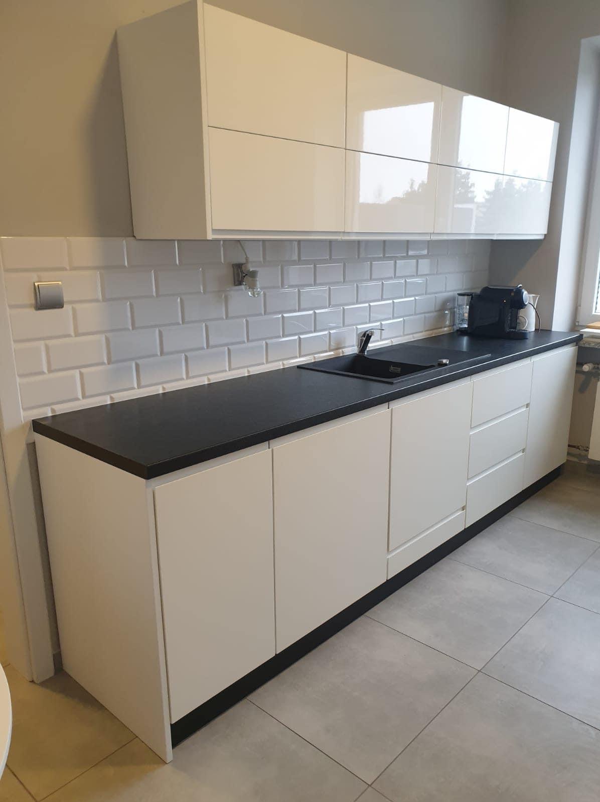 Biała, nowoczesna zabudowa kuchenna z czarnym blatem, płytki typu metro na ścianie, widoczny ekspres do kawy.