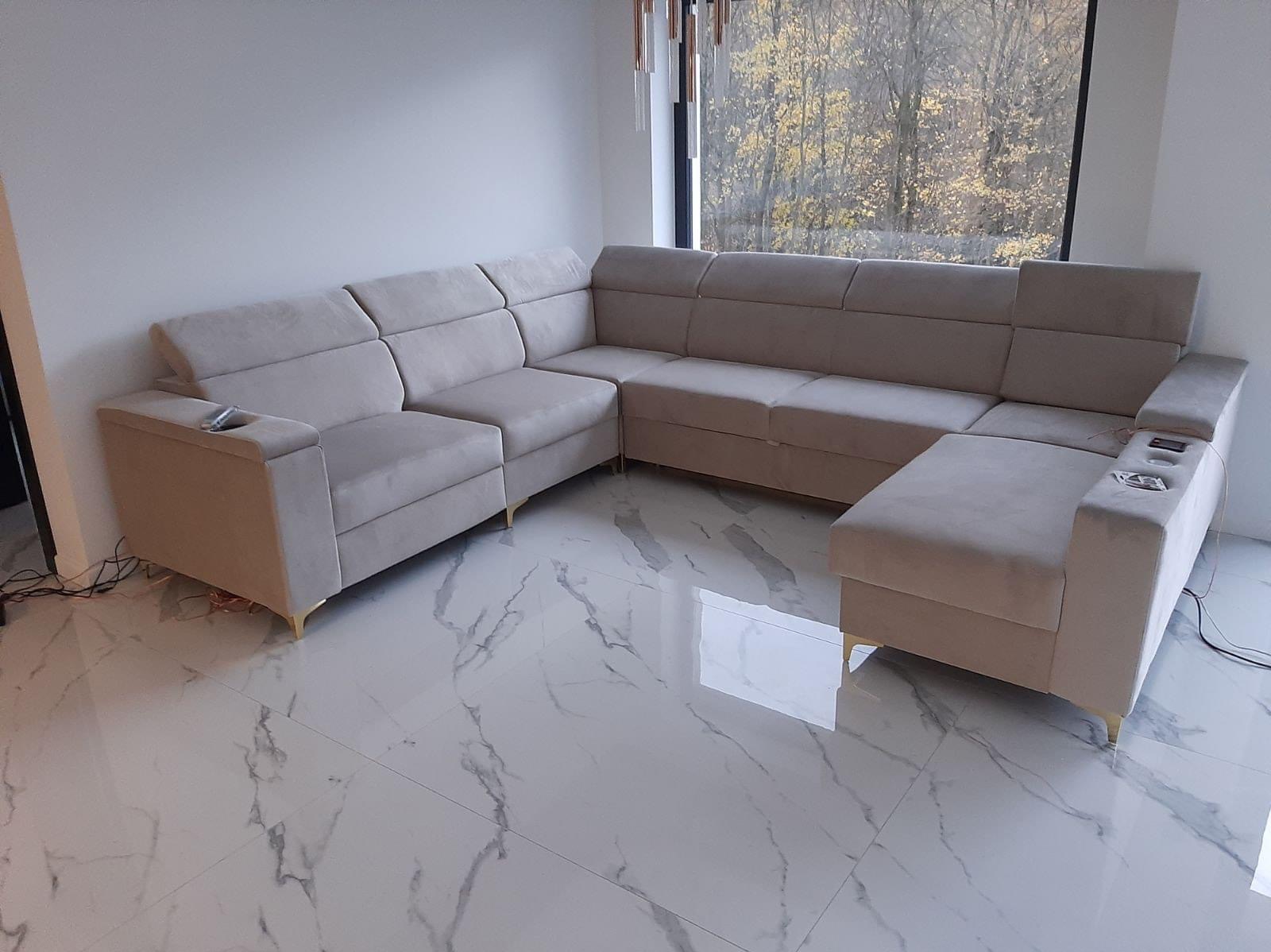 Beżowy, modułowy narożnik z tapicerką welurową, złotymi nóżkami i wbudowanym uchwytem na napoje, ustawiony na marmurowej podłodze w nowoczesnym salonie z dużym oknem.