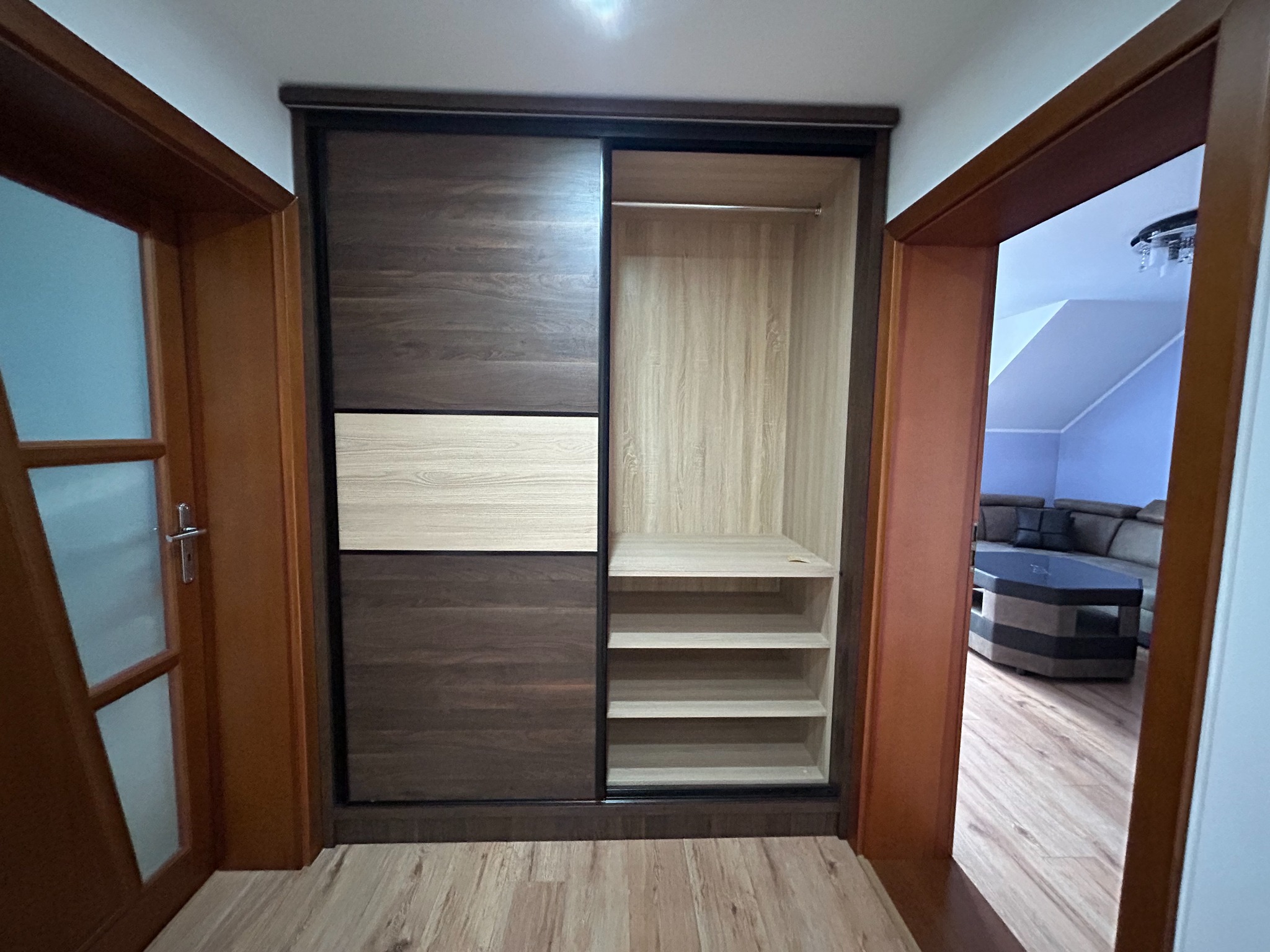 Szafa wnękowa z przesuwnymi drzwiami w kolorze drewna, z półkami i drążkiem, widok z korytarza na salon.