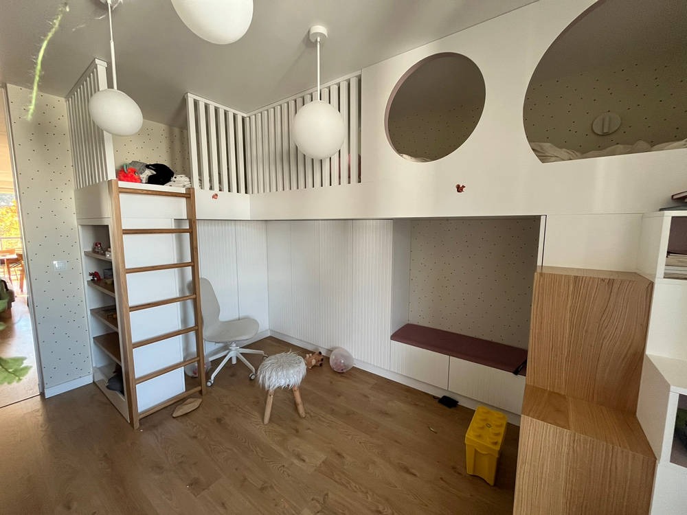 Pokój dziecięcy z zabudową na wymiar: antresola z barierką, schody z półkami, białe ściany z okrągłymi oknami, drewniana podłoga, oświetlenie punktowe, krzesło na kółkach i pufa.