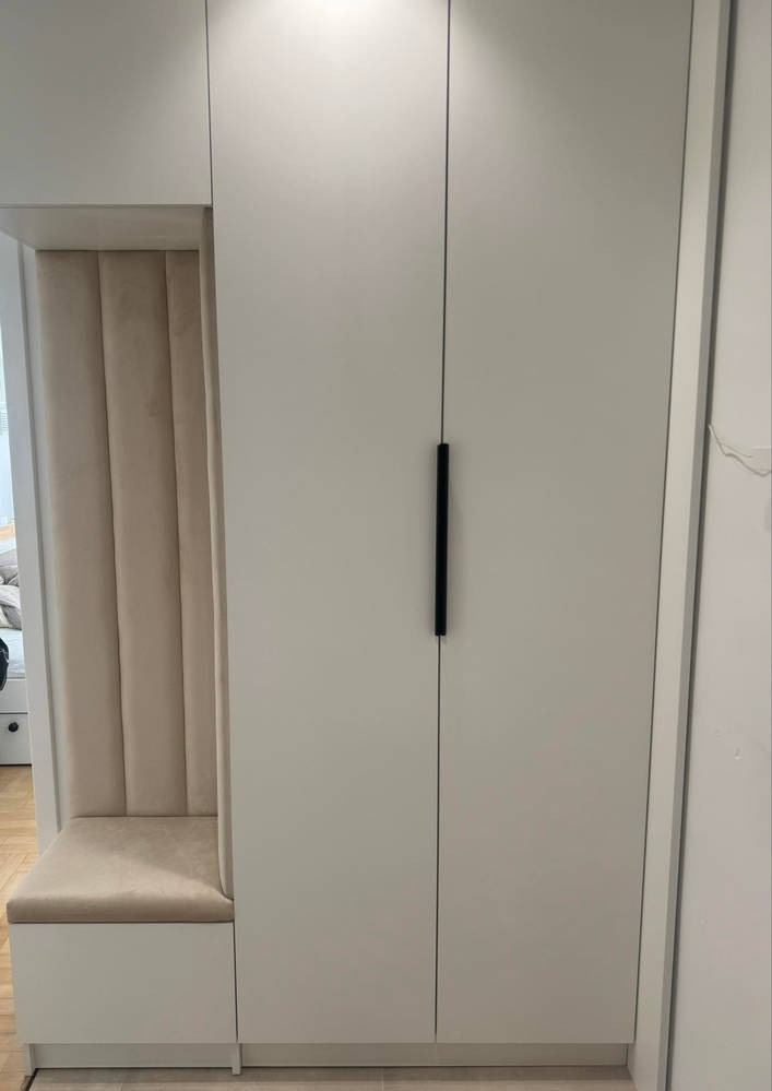 Biała szafa wnękowa z siedziskiem i tapicerowanym panelem ściennym w beżowym odcieniu, minimalistyczne czarne uchwyty.