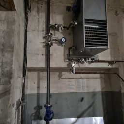 Gold-welder - Szara nagrzewnica przemysłowa z wentylatorem i rurami, zamontowana na betonowej ścianie z widocznymi instalacjami hydraulicznymi i elektrycznymi.