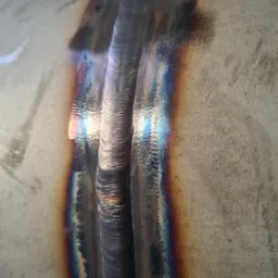 Zbliżenie na świeży spaw na metalu, widoczne tęczowe przebarwienia termiczne i struktura spoiny.