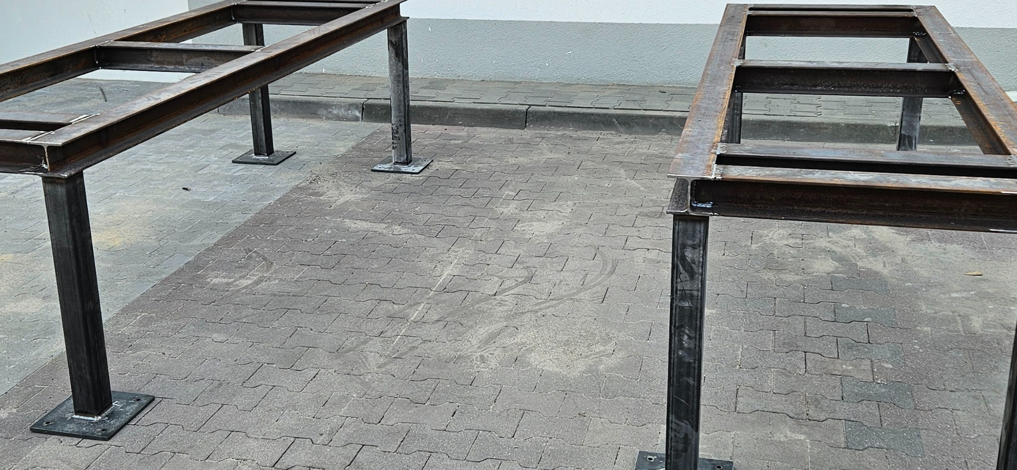 Dwie surowe, metalowe konstrukcje ramowe z profili stalowych, stojące na kostce brukowej, prawdopodobnie przygotowane do dalszej obróbki lub montażu.