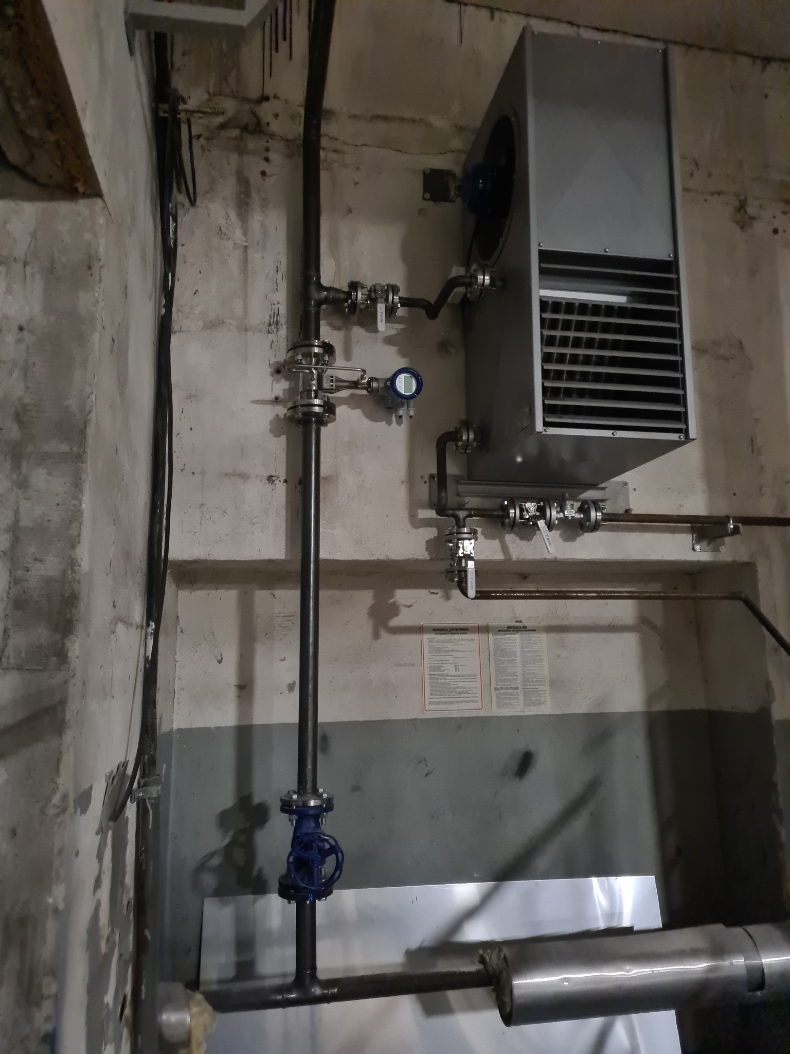 Szara nagrzewnica przemysłowa z wentylatorem i rurami, zamontowana na betonowej ścianie z widocznymi instalacjami hydraulicznymi i elektrycznymi.