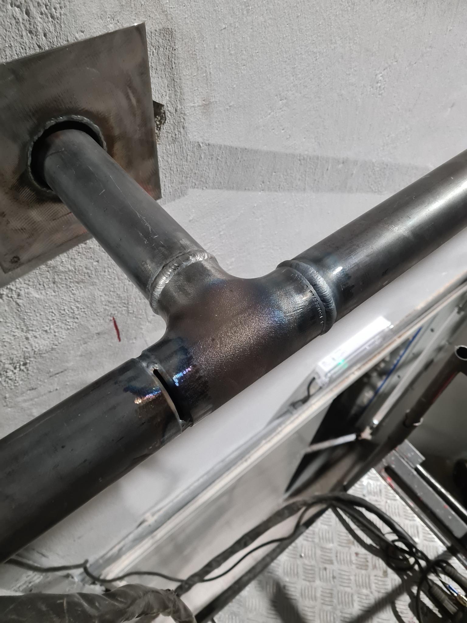 Metalowa rura spawana trójnikiem, przymocowana do ściany za pomocą kwadratowej blachy, widoczne ślady spawania i niewielkie uszkodzenie na jednej z rur.