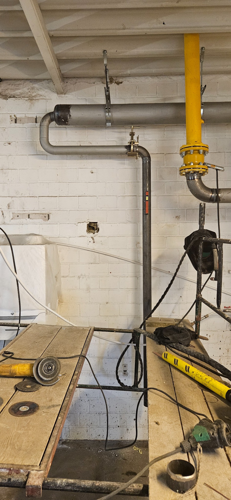Wnętrze z instalacją rur, widoczne narzędzia ślusarskie na drewnianym stole, biała ceglana ściana w tle. Żółte rury kontrastują z szarymi.