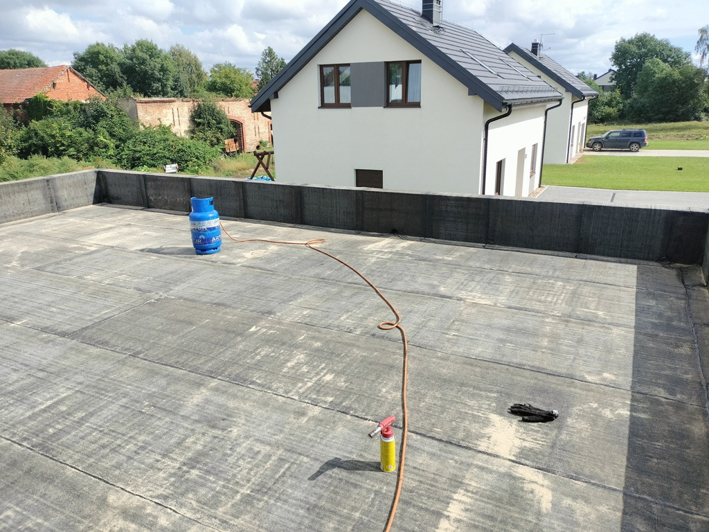 Płaski dach pokryty papą termozgrzewalną, widoczny palnik dekarski, butla gazowa oraz rękawice robocze, w tle nowoczesny dom jednorodzinny z szarym dachem.