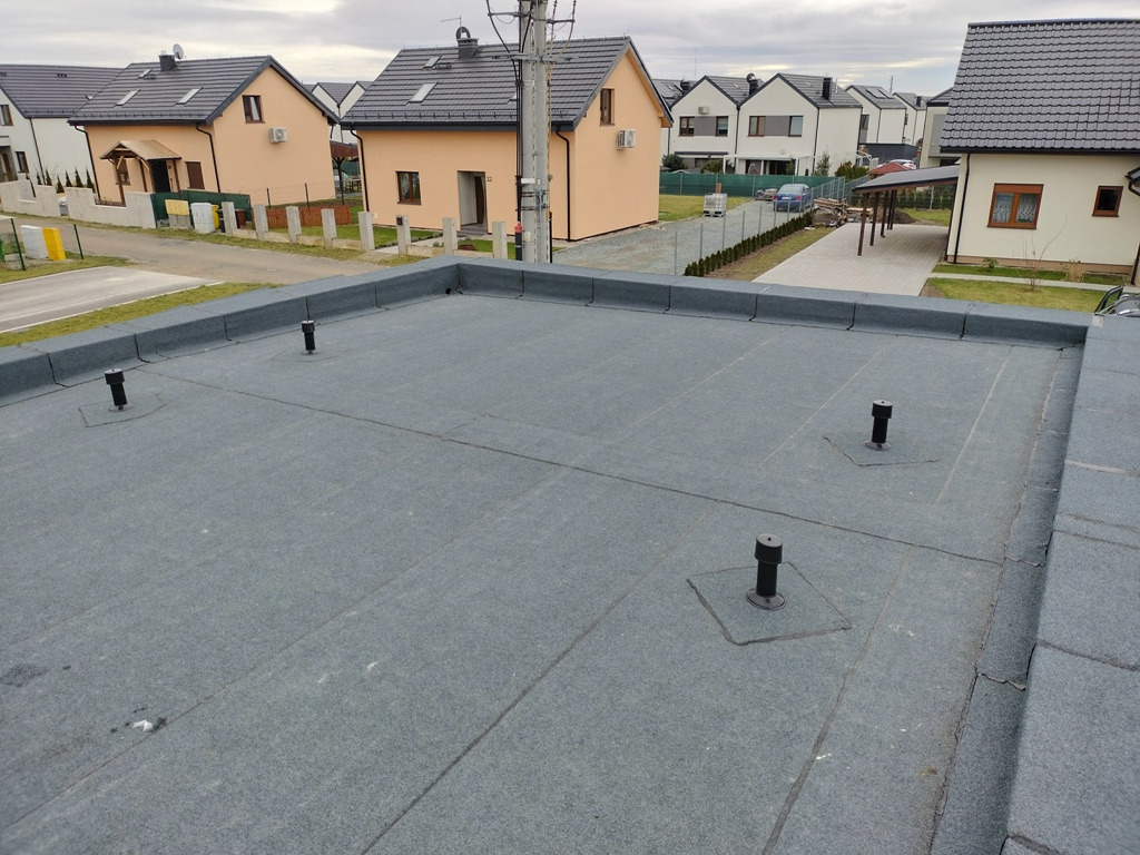 Płaski dach pokryty papą termozgrzewalną z widocznymi kominkami wentylacyjnymi i obróbkami blacharskimi na tle zabudowy jednorodzinnej.