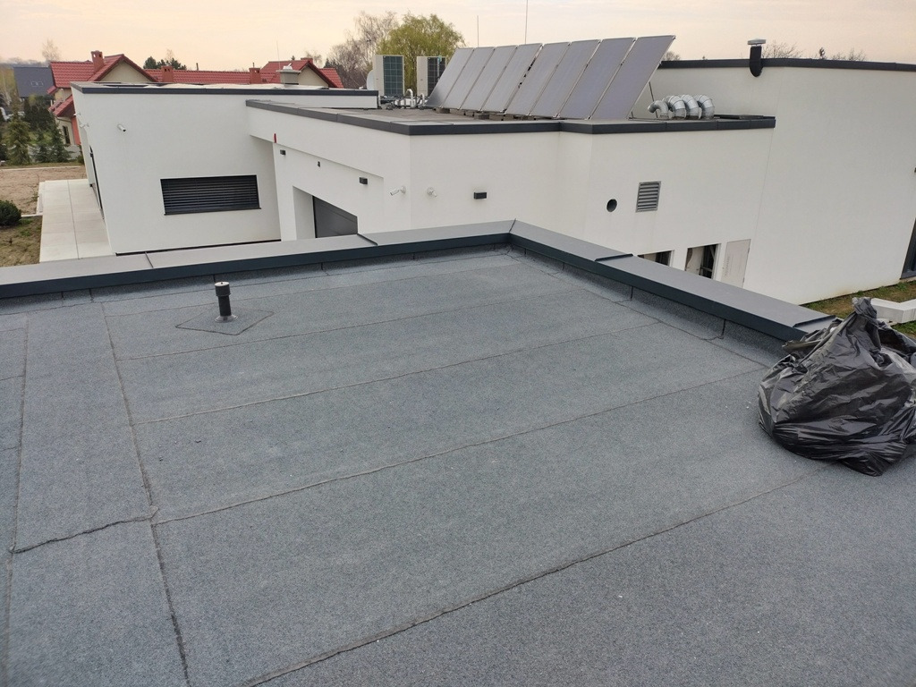 Płaski dach pokryty papą termozgrzewalną na nowoczesnym budynku z panelami solarnymi i workiem na odpady w rogu.