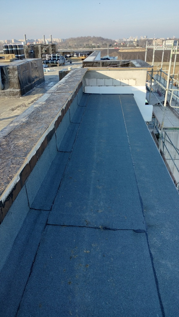 Widok z góry na płaski dach pokryty papą termozgrzewalną, widoczne łączenia, obróbki blacharskie i elementy konstrukcyjne dachu, w tle panorama miasta.