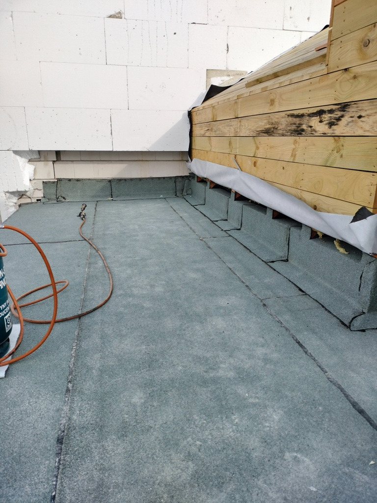 Wykończona płaska powierzchnia dachu pokryta papą termozgrzewalną, widoczne łączenia, obróbki przy ścianie z bloczków betonowych i drewnianej konstrukcji, butla gazowa i palnik dekarski w lewym...