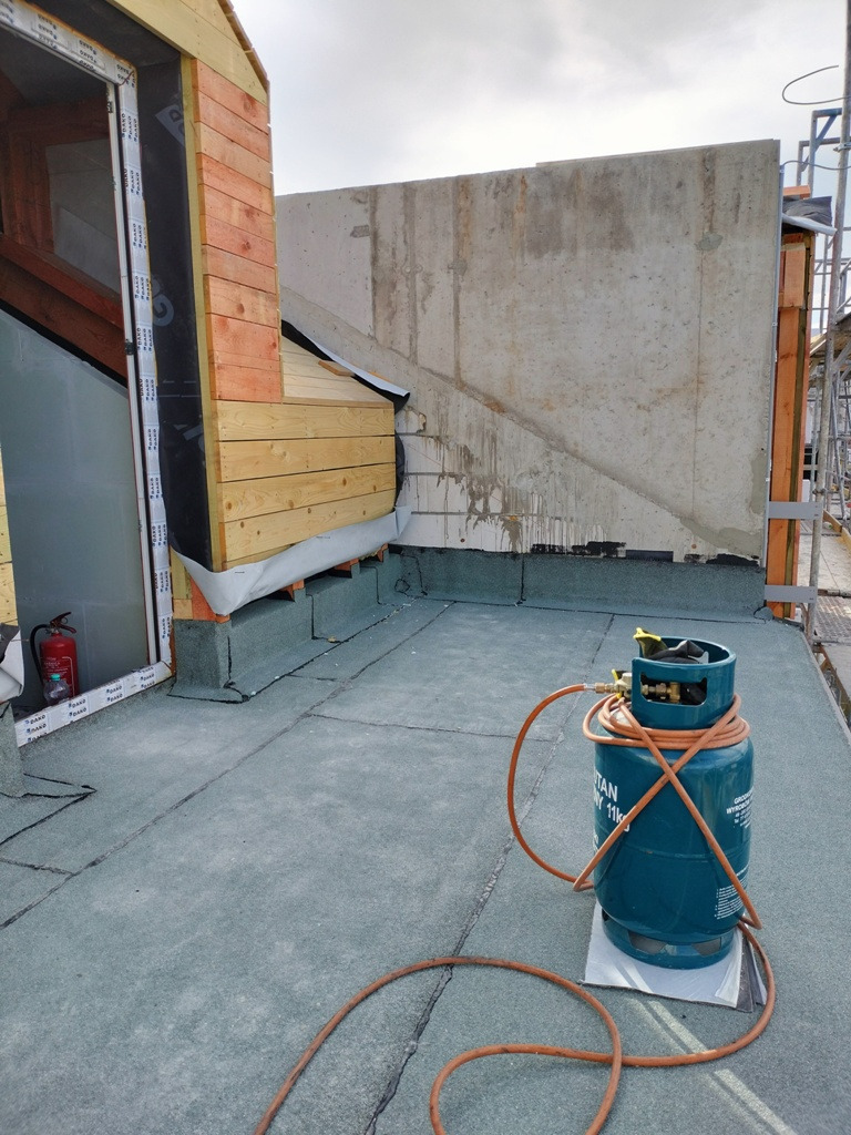 Płaski dach pokryty papą termozgrzewalną z widoczną butlą gazową i palnikiem, fragment drewnianej konstrukcji i ściana budynku w tle.