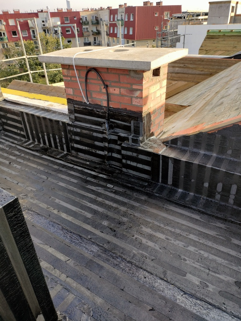 Uszczelniony komin ceglany z betonową czapą na dachu pokrytym papą termozgrzewalną, widoczne detale izolacji i obróbki blacharskiej u podstawy komina.