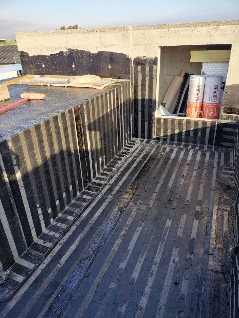 Przygotowanie podłoża na dachu płaskim z izolacją bitumiczną w pasach, widoczne rolki izolacji Bauder i narzędzia pracy.