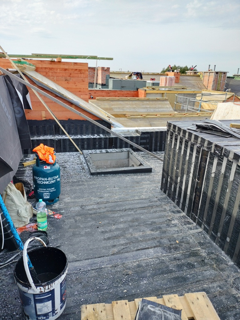 Widok na dach w trakcie prac dekarskich, widoczna butla gazowa, wiadro z substancją, materiały izolacyjne i fragmenty konstrukcji dachu z cegły.