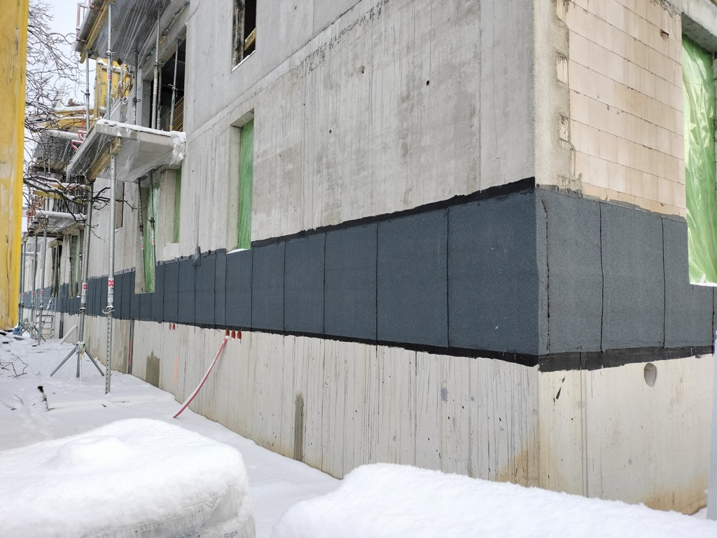 Izolacja fundamentów budynku w trakcie budowy, widoczne szare panele izolacyjne przytwierdzone do betonowej ściany fundamentowej, śnieg na ziemi.