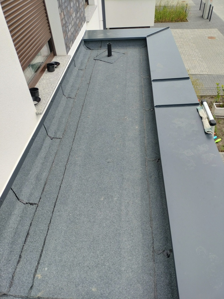 Płaski dach pokryty papą z widocznymi obróbkami blacharskimi, kominkiem wentylacyjnym i fragmentem elewacji budynku z roletami zewnętrznymi.