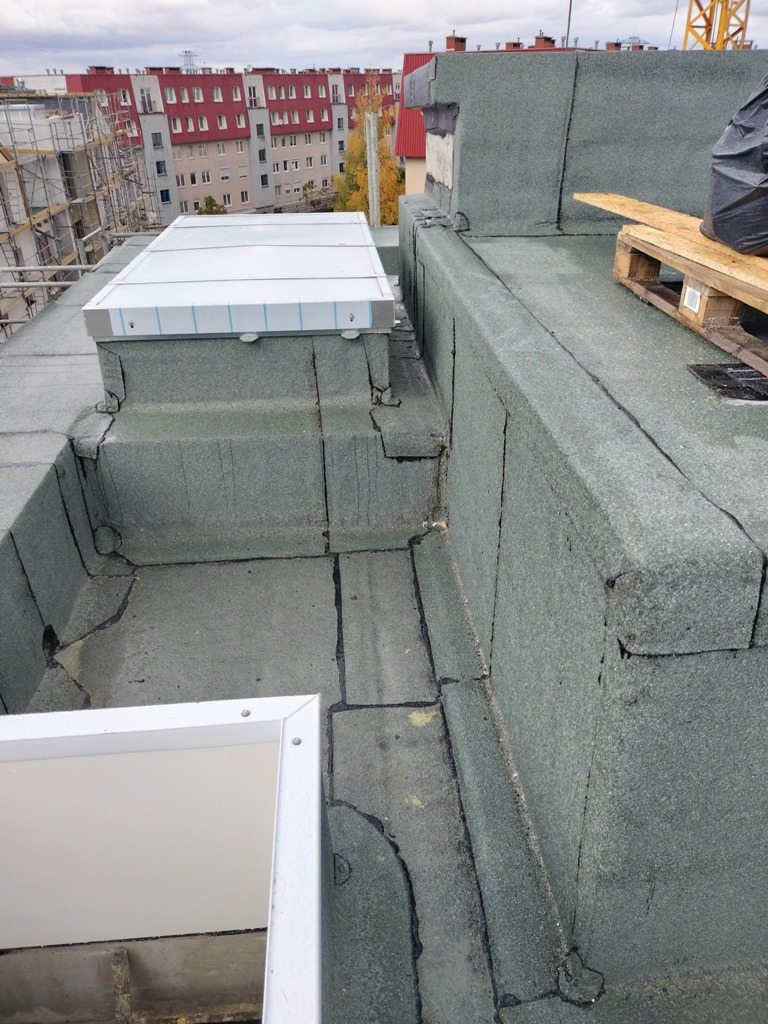Płaski dach pokryty papą z wbudowanymi świetlikami dachowymi, widoczne łączenia papy i detale obróbki, w tle budynki mieszkalne i dźwig budowlany.