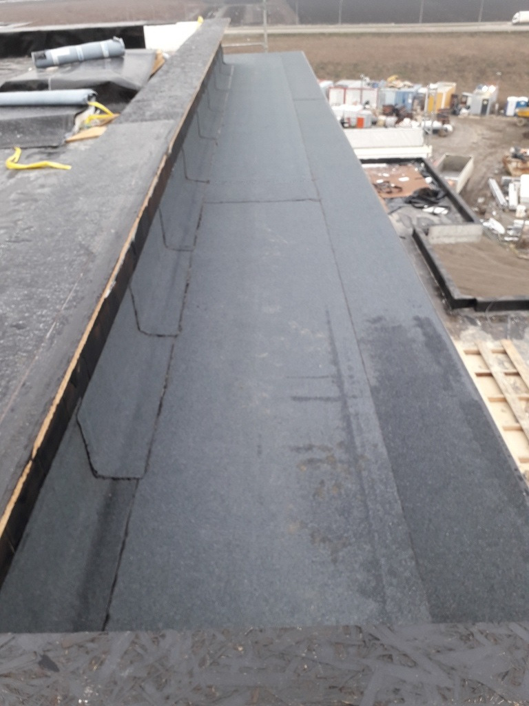 Fragment płaskiego dachu pokrytego papą termozgrzewalną, widoczne łączenia i elementy wykończeniowe, w tle zabudowania przemysłowe.