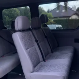 Wnętrze srebrnego busa z trzema rzędami siedzeń z szarymi tapicerkami w geometryczne wzory, widok z otwartych tylnych drzwi na dom z brązowym dachem w tle.