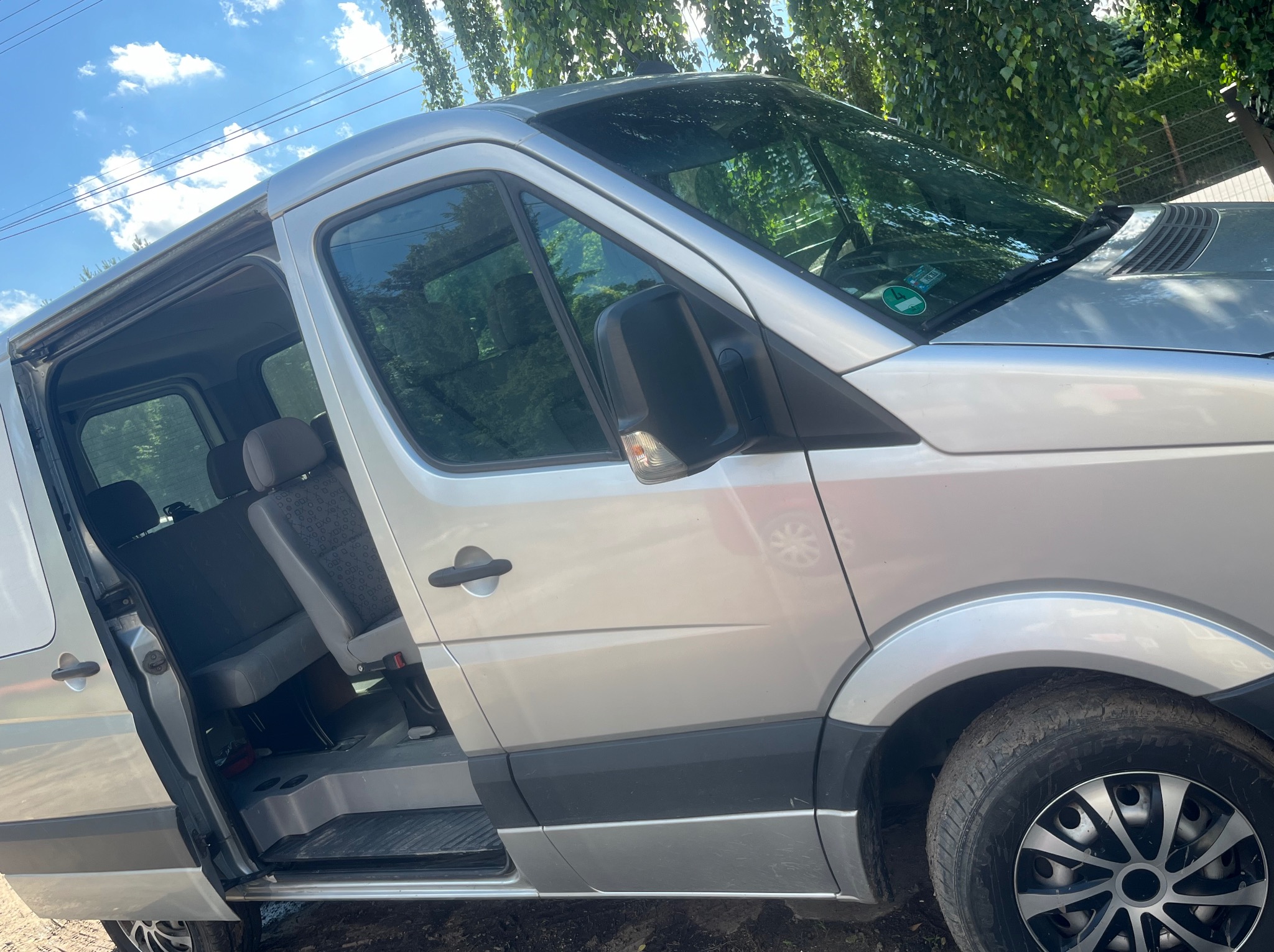 Srebrny van z otwartymi drzwiami bocznymi, widoczne fotele pasażerskie wewnątrz, na zewnątrz drzewa i błękitne niebo.