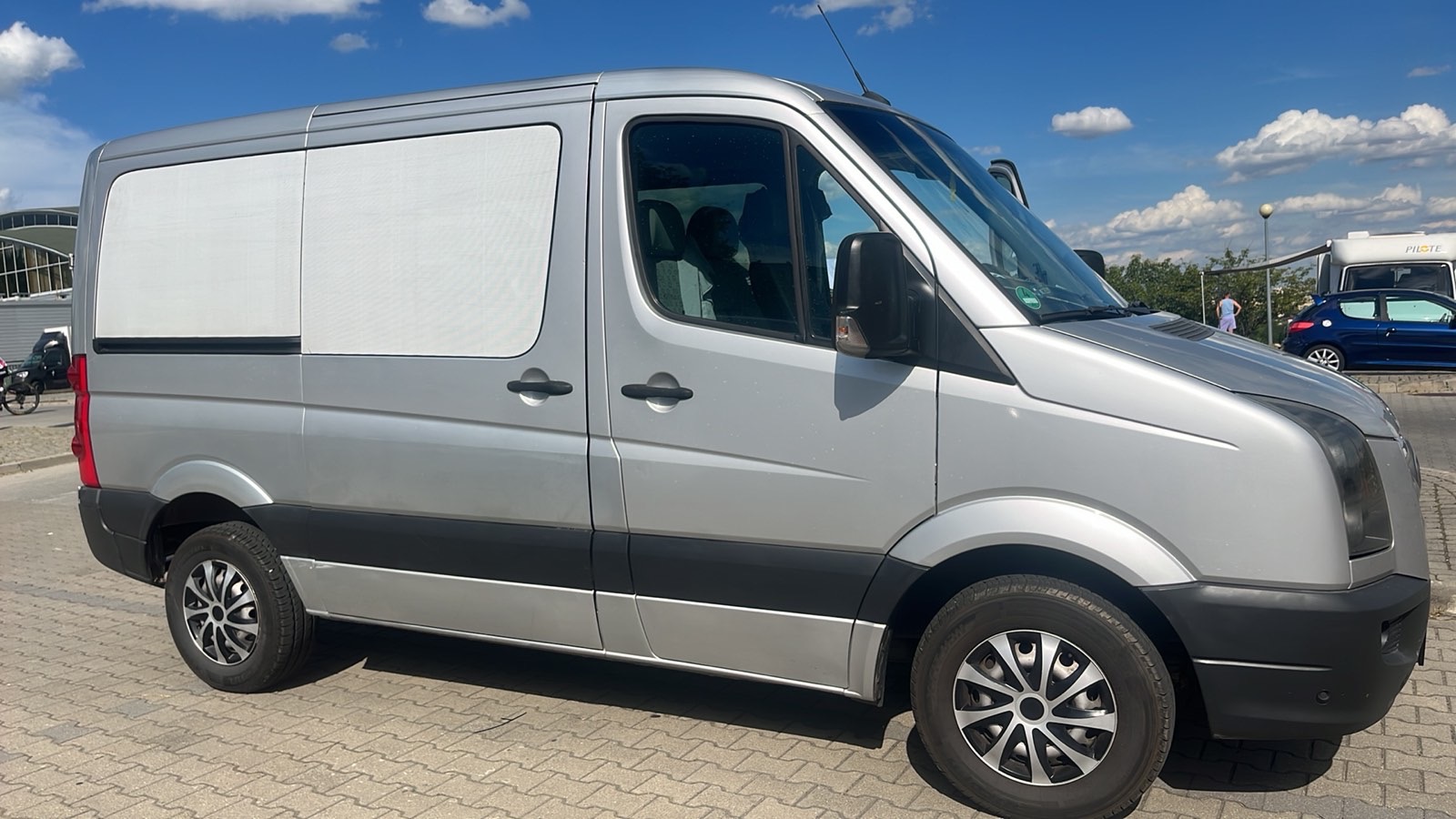 Srebrny van z zasłoniętymi szybami z tyłu, zaparkowany na kostce brukowej, z widocznym fragmentem nieba i innymi pojazdami w tle.