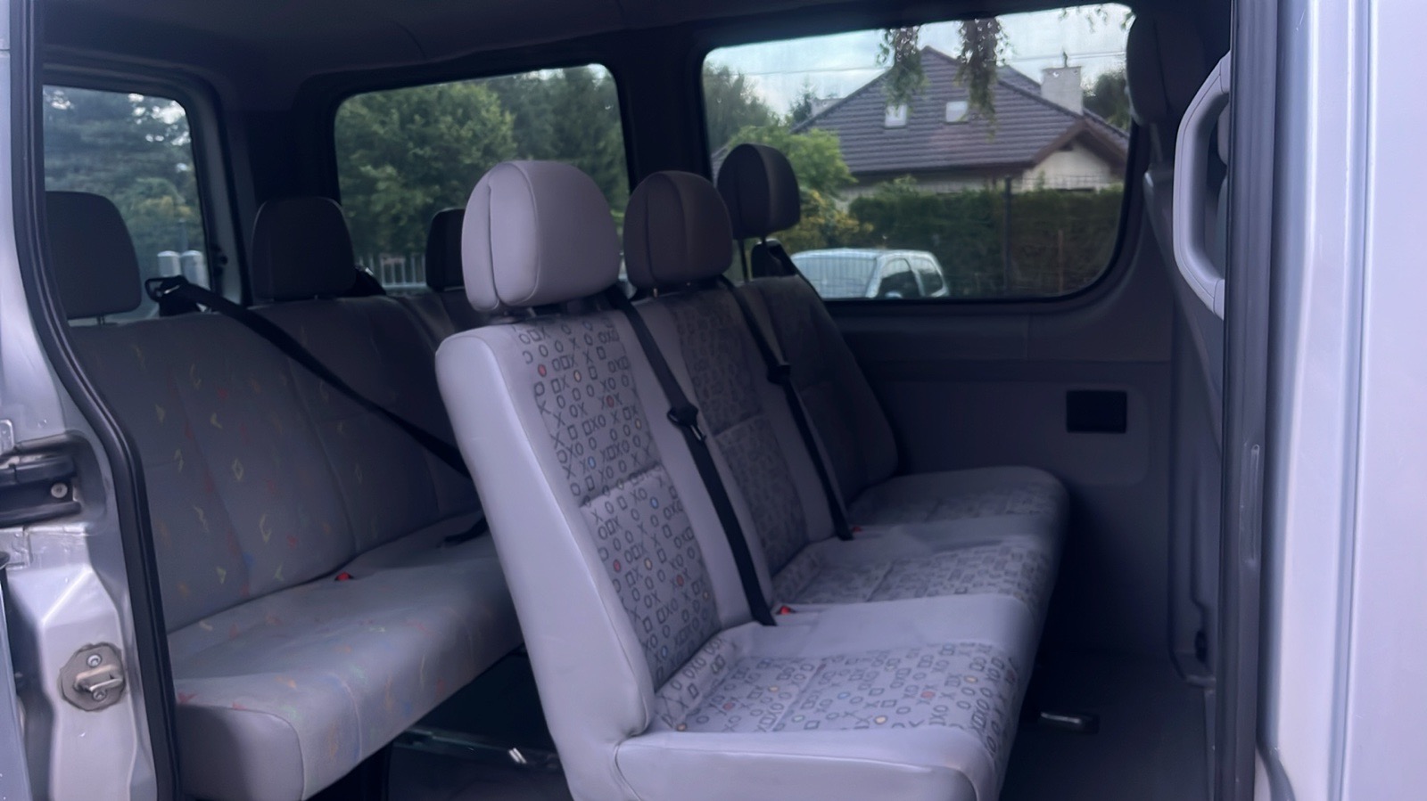 Wnętrze srebrnego busa z trzema rzędami siedzeń z szarymi tapicerkami w geometryczne wzory, widok z otwartych tylnych drzwi na dom z brązowym dachem w tle.