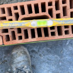 Budownictwo i AutoSerwis Piotr Grajek - Cegła dziurawka poziomowana żółtą poziomicą, widoczny fragment buta roboczego murarza i młotek