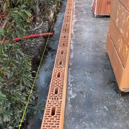 Budownictwo i AutoSerwis Piotr Grajek - Wąski wykop z ułożonym pierwszym rzędem bloczków ceramicznych z pustakami, wyznaczony żółtą linką, przygotowany pod fundament budynku.