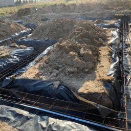 Budownictwo i AutoSerwis Piotr Grajek - Przygotowanie wykopu pod fundamenty z izolacją folią i zbrojeniem stalowym, widoczny nasyp ziemi i kamieni.