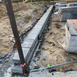 Budownictwo i AutoSerwis Piotr Grajek - Wznoszenie fundamentu z betonowych bloczków z użyciem stalowego słupka i sznurka murarskiego, widoczna izolacja przeciwwilgociowa i zapas bloczków na budowie.