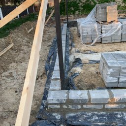 Budownictwo i AutoSerwis Piotr Grajek - Wznoszenie fundamentów z bloczków betonowych, widoczne szalunki z desek, czarna folia izolacyjna i palety z zapakowanymi bloczkami w tle.