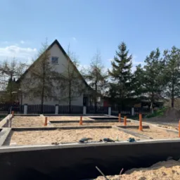 Wylane fundamenty domu jednorodzinnego z widoczną hydroizolacją, piaskiem wewnątrz i wystającymi rurami kanalizacyjnymi, na tle domu z ogrodem w słoneczny dzień.