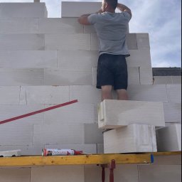 Budownictwo i AutoSerwis Piotr Grajek - Murarz w szarej koszulce i czarnych spodenkach układa białe bloczki z betonu komórkowego na budowie, stojąc na rusztowaniu z poziomnicą i tubą kleju Dryfix extra, widok z dołu na niebo.