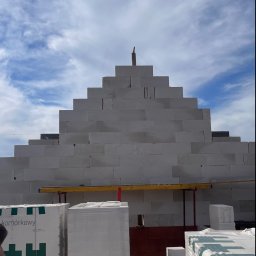 Budownictwo i AutoSerwis Piotr Grajek - Wznoszona ściana z białych bloczków komórkowych o piramidalnym kształcie, z widocznym poziomowaniem i paletami materiałów budowlanych u podstawy, na tle błękitnego nieba.