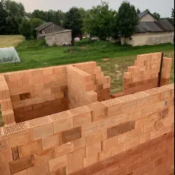 Wznoszenie ścian domu z czerwonych bloczków ceramicznych Porotherm na wsi, widoczne fragmenty muru z otworami, w tle zabudowania gospodarcze i zieleń.